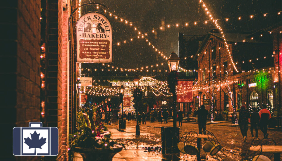 VisitorsInsurance.ca A snowy night scene at the Distillery Winter Village in Toronto, featuring festive string lights hanging over cobblestone streets and historic red brick buildings, including the Brick Street Bakery.