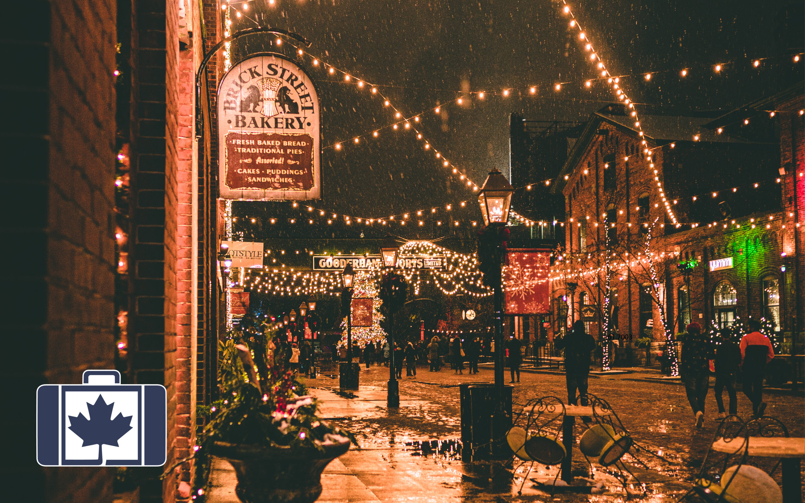 VisitorsInsurance.ca A snowy night scene at the Distillery Winter Village in Toronto, featuring festive string lights hanging over cobblestone streets and historic red brick buildings, including the Brick Street Bakery.