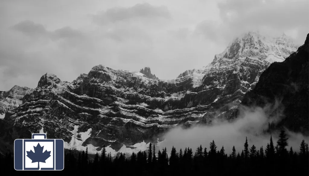 Adventure Sports in Canada Are You Covered A black and white image of a landscape of the snow-dusted Rocky Mountains in Alberta, featuring prominent horizontal rock layers, low-lying fog over a dark pine forest silhouette.
